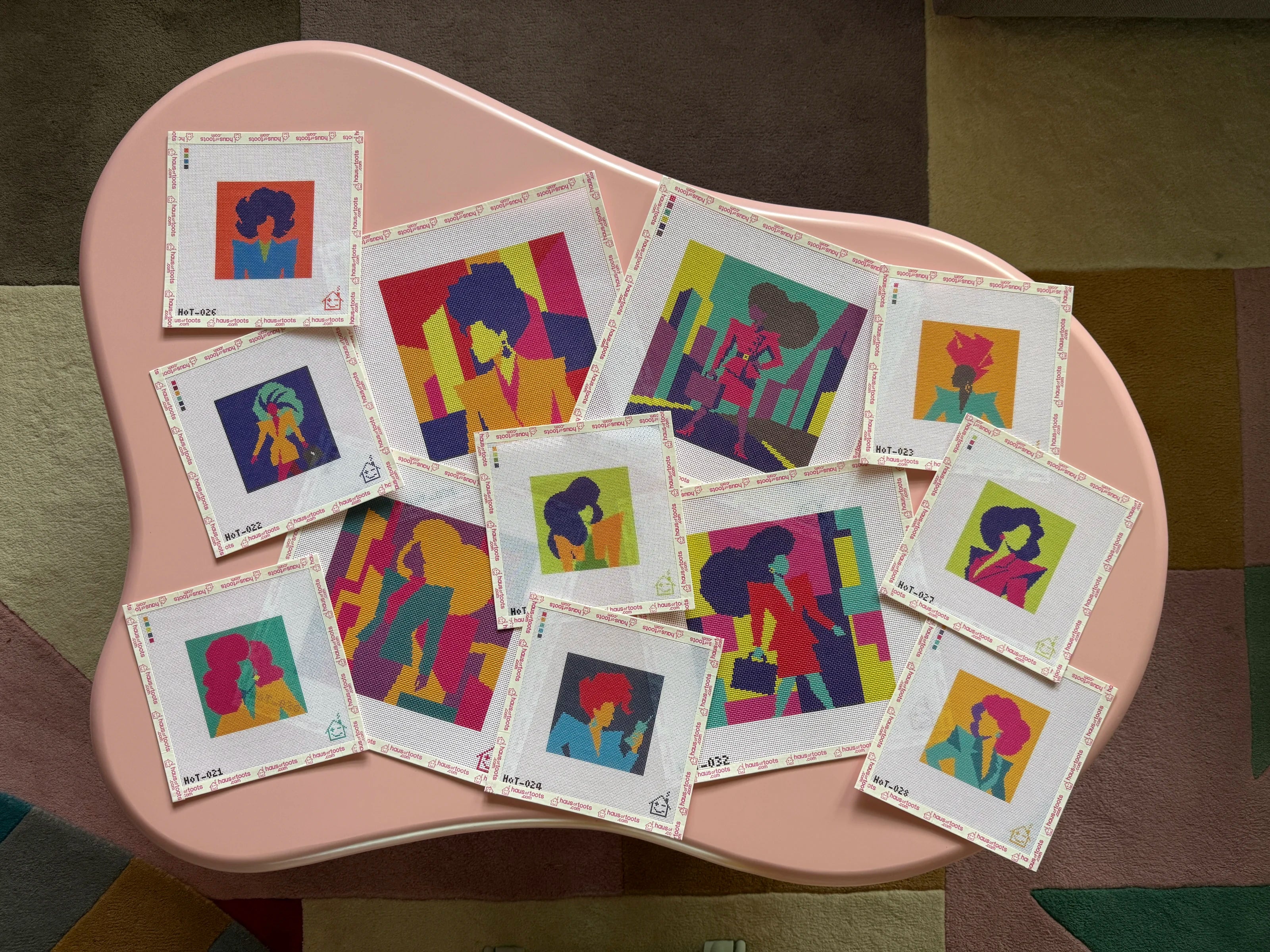 Twelve colorful Main Stage needlepoint canvases arranged on a blush tray.